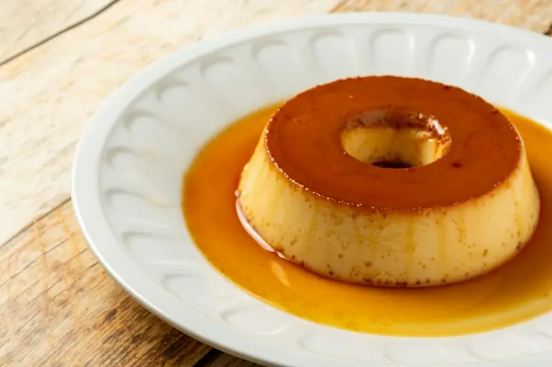 Pudim de leite condensado com furo no meio, coberto de calda de caramelo, num prato branco sobre mesa de madeira