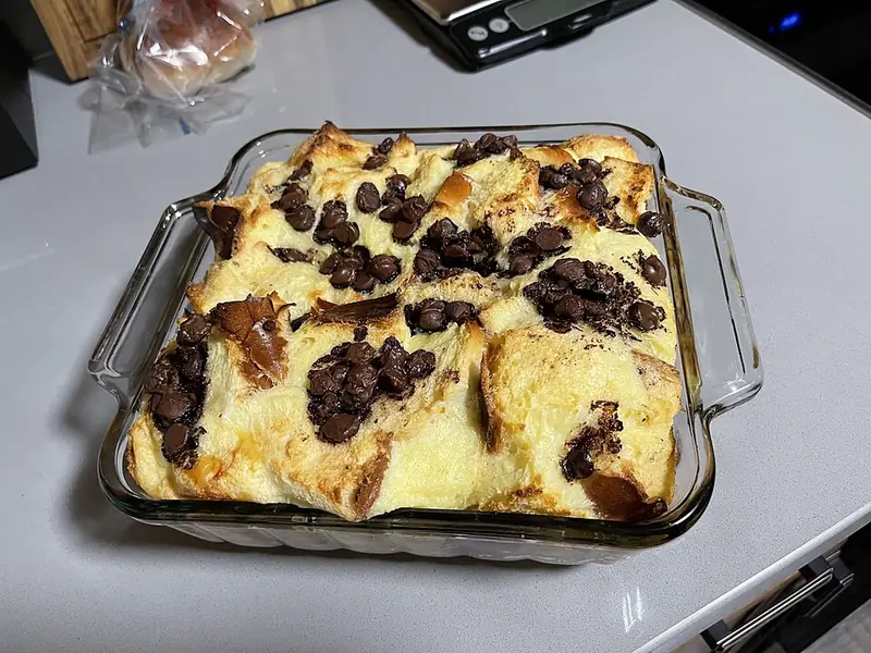 Pudim de pão assado numa travessa de vidro, com pedaços de pão dourados e gotas de chocolate